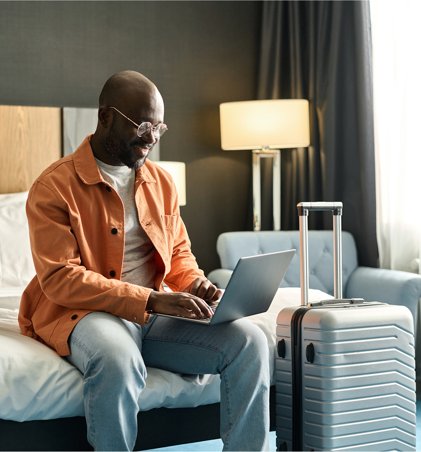 Man using laptop beside suitcase on bed.